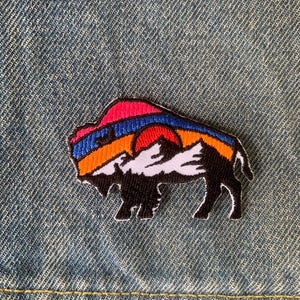 Op de afbeelding: Geborduurde patch met een silhouet van een bizon met een zonsondergangontwerp. De patch heeft een zwarte bizon met een rode, oranje, blauwe en witte zonsondergang. De patch bevindt zich op een denim achtergrond.