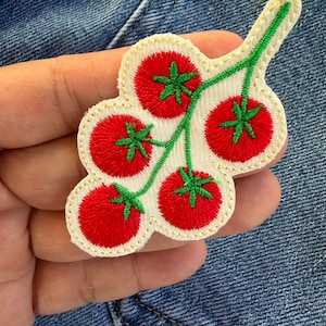 以下が含まれることがあります： 緑色の茎と葉が付いた、5つの赤いトマトの房が刺繍されたパッチ。トマトは赤い糸で、茎と葉は緑色で縫われています。パッチは白い背景とクリーム色の縁取りが施されています。