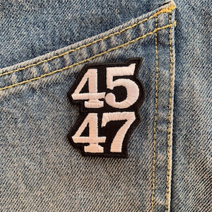 以下が含まれることがあります： 黒い背景に白い刺繍で45と47の数字が書かれたパッチ。パッチは青いデニムのポケットに縫い付けられている。