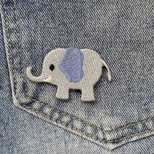 Op de afbeelding: Een geborduurde grijze olifant patch met een paars oor, genaaid op een blauwe denim zak.
