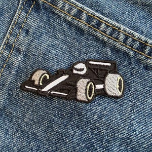Peut inclure: Un écusson brodé représentant une voiture de course noire avec des accents blancs et des roues grises. Le motif inclut un petit casque blanc visible au niveau du cockpit. L'écusson est fixé sur un tissu en jean bleu avec des coutures apparentes.