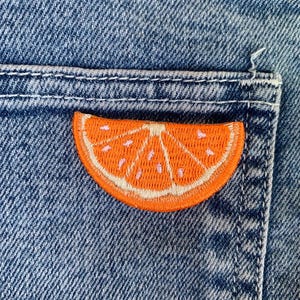 Może przedstawiać: Zbliżenie na kieszeń dżinsową z naszywką w kształcie półkola cytrusowego, wyszywaną na pomarańczowo.