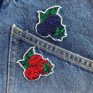 以下が含まれることがあります： 青いデニムの背景に、2つの刺繍されたアイロン接着パッチ。1つのパッチは赤いラズベリーの房、もう1つは青いブルーベリーの房を特徴としています。