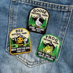 May include: Three embroidered patches on blue denim fabric. One patch features a goose with the text GOOSE NATIONAL PARK HONK AROUND FIND OUT. Another shows a bee with BEE NATIONAL PARK MESS WITH THE HIVE FIND OUT. The third shows a frog with FROG NATIONAL PARK HOP IN, CHILL OUT. LEAVE NO DRAMA.
