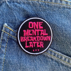 Può includere: Una toppa rotonda nera con la scritta "ONE MENTAL BREAKDOWN LATER" in lettere rosa acceso. La toppa ha un bordo bianco ed è posta su uno sfondo di denim blu.