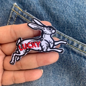 Op de afbeelding: Een wit en zwart geborduurd embleem in de vorm van een rennend konijn met het woord "LUCKY" in rood. De patch bevindt zich op een denim achtergrond.