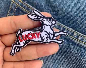 May include: A white and black embroidered patch in the shape of a running rabbit with the word "LUCKY" in red. The patch is on a denim background.