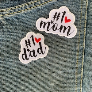 May include: Two white embroidered patches with black text and red hearts. One patch says "#1 mom" and the other says "#1 dad". The patches are on a denim background.