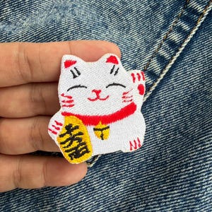 May include: White embroidered Maneki-neko cat patch with red accents on the ears, collar, and paws. The cat holds a gold coin with black Japanese characters. The patch is on a blue denim background.