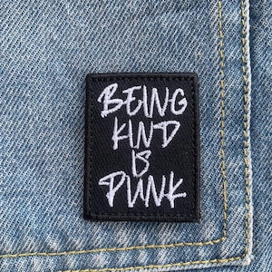 Puede incluir: Un parche cuadrado negro con las palabras "BEING KIND IS PUNK" bordadas en blanco, sobre un fondo de mezclilla azul. El parche está cosido a la mezclilla con hilo amarillo.