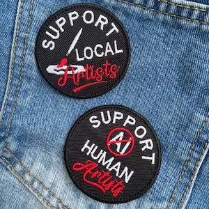 May include: Two circular black embroidered patches with white and red text. The top patch reads SUPPORT LOCAL Artists with a paintbrush icon. The bottom patch reads SUPPORT HUMAN Artists with a red crossed-out AI symbol in the center.