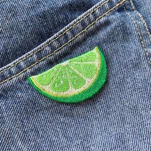 以下が含まれることがあります： 青いデニムの背景に、緑色の刺繍されたライムのくさび形のワッペンが貼られています