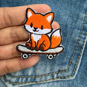 May include: An embroidered patch featuring a cartoon fox with orange and white fur sitting on a light grey skateboard. The fox has dark brown paws, pointed ears, and a bushy tail. The design is outlined in black thread with a clean, stitched finish.