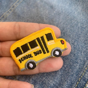 以下が含まれることがあります： 黒い窓とタイヤ、白い「SCHOOL BUS」の文字が刺繍された黄色のアイロン接着パッチ。スクールバスのデザインです。
