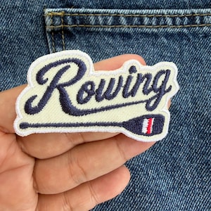 May include: An embroidered patch featuring the word Rowing in dark blue cursive script over a cream-coloured background. The design includes a dark blue oar graphic underneath the text with a small red and white stripe detail on the oar blade.