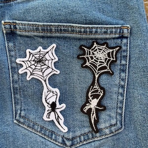 Pode incluir: Dois patches bordados com um desenho de teia de aranha e aranha. Um é branco com detalhes em preto, o outro preto com detalhes em branco. Estão presos ao bolso de trás de uns jeans azuis claros.