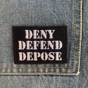 May include: Black patch with white embroidered text that reads "DENY DEFEND DEPOSE" on a blue denim background.