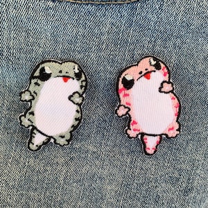 Może przedstawiać: Dwa haftowane naszywki z kreskówkowymi gekonami. Gekon po lewej stronie jest zielony i biały, a gekon po prawej stronie jest różowy i biały.