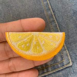 Könnte beinhalten: Gestickter Aufnäher in Form einer Zitronenscheibe. Der Aufnäher hat ein gelbes Inneres mit weißen Akzenten und einen orangefarbenen Rand. Er ist wahrscheinlich für Kleidung oder Accessoires bestimmt.