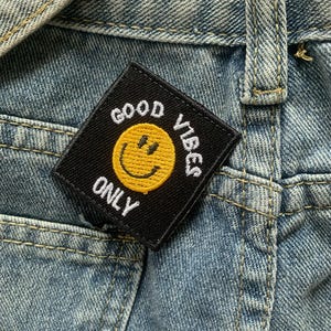 Może przedstawiać: Czarny kwadratowy patch z białym tekstem "Good Vibes Only" i żółtą buźką w środku. Patch jest przyszyty do niebieskiego dżinsu.