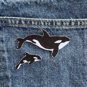 May include: Two embroidered patches featuring orca whales in black and white thread. The larger orca is positioned above the smaller orca, both displayed against a textured blue denim fabric background with visible gold stitching along the seams.