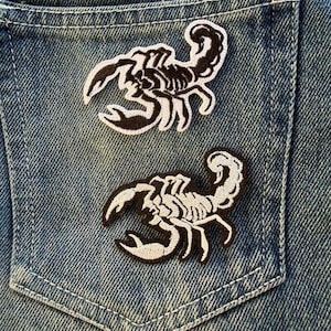 以下が含まれることがあります： 黒と白のサソリの刺繍パッチが2つ、デニムのポケットに付いています。パッチは、湾曲した尾と爪のあるハサミを持つ詳細なサソリのデザインが特徴です。