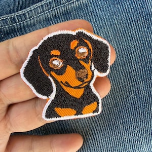 Op de afbeelding: Geborduurde patch met een teckel hond met zwarte, oranje en witte details. De patch heeft een witte rand en is ontworpen om op stof te strijken of te naaien. Het gezicht van de hond is gedetailleerd met bruin en oranje.