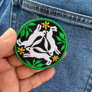 Może przedstawiać: Okrągła, haftowana naszywka z trzema białymi królikami na czarnym tle, otoczona zielonymi liśćmi i żółtymi kwiatami. Naszywka ma zieloną obwódkę i jest umieszczona na tle niebieskiego dżinsu.