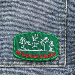 Könnte beinhalten: Grüner gestickter Aufnäher mit weißem Blumendesign und dem Text "NO RAIN NO FLOWERS" in Rot. Der Aufnäher ist rechteckig mit abgerundeten Ecken und auf Denim-Stoff genäht.