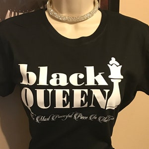 May include: Black t-shirt with the words "black QUEEN" in large white letters and a white chess queen graphic. Below the text is the phrase "The Most Powerful Piece In The Game" in white script. A rhinestone choker necklace is around the mannequin's neck.