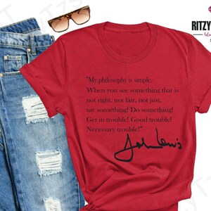 May include: A red t-shirt featuring a quote by John Lewis and his signature, alongside blue jeans and sunglasses. The shirt's text reads "My philosophy is simple..." and is part of a Black Culture collection.
