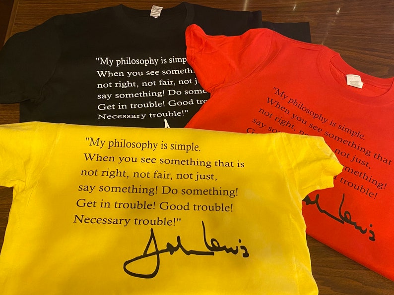 May include: Four t-shirts in black, red, and yellow are displayed. Each shirt features the quote "My philosophy is simple...say something!" and a signature. The shirts are made of a soft material.