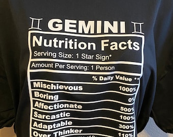 May include: Black t-shirt featuring a white graphic in the style of a nutrition facts label, listing Gemini traits such as "Mischievous" and "Adaptable". The text includes percentages. A sparkly rhinestone necklace is visible at the neckline.