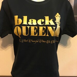 May include: Black t-shirt with gold lettering that reads "black QUEEN" and a gold chess queen graphic. Below the text is the phrase "The Most Powerful Piece In The Game". A silver necklace is around the neck of the mannequin.