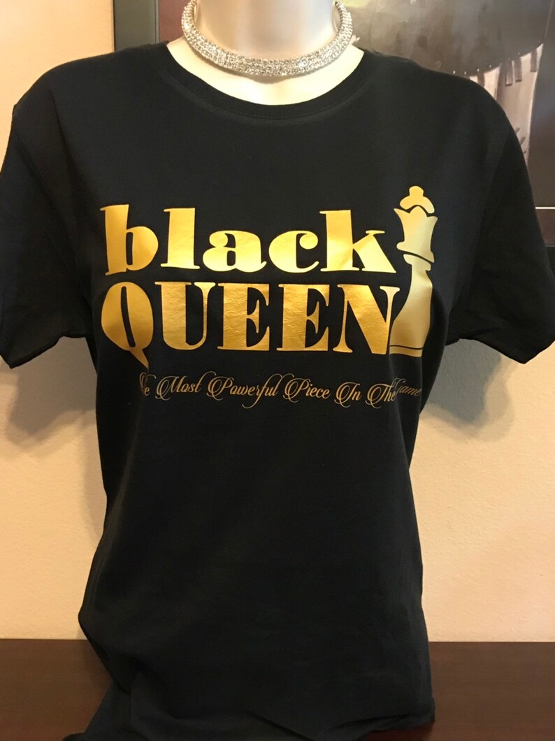 May include: Black t-shirt with gold lettering that reads "black QUEEN" and a gold chess queen graphic. Below the text is the phrase "The Most Powerful Piece In The Game". A silver necklace is around the neck of the mannequin.