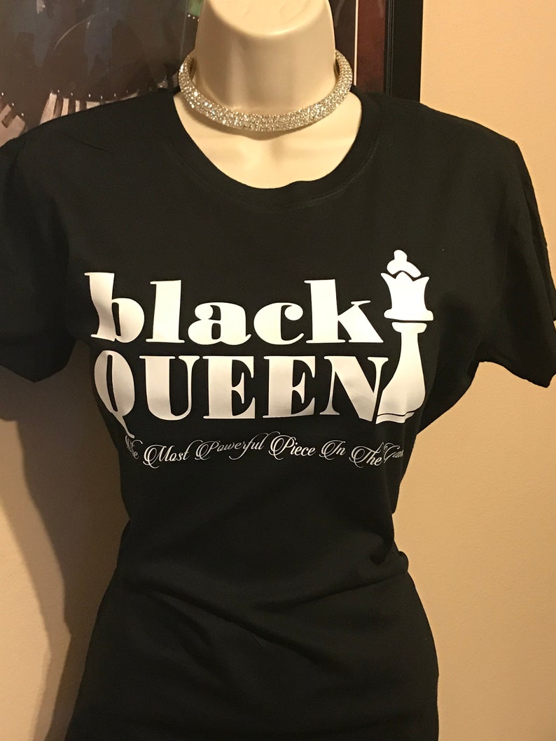 May include: Black t-shirt with the words "black QUEEN" in large white letters and a white chess queen graphic. Below the text is the phrase "The Most Powerful Piece In The Game" in white script. A rhinestone choker necklace is around the mannequin's neck.