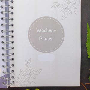 May include: A gray and white weekly planner with a circle on the front that says "Wochen-Planer" in a gray font.
