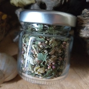 May include: A small glass jar with a silver lid filled with dried herbs and flowers. The jar is sitting on a wooden surface.