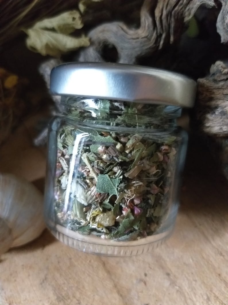 May include: A small glass jar with a silver lid filled with dried herbs and flowers. The jar is sitting on a wooden surface.
