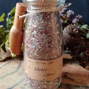 Könnte beinhalten: Ein Glasgefäß mit einem Korkverschluss, gefüllt mit einer Mischung aus Kräutern, Blumen und Kristallen. Das Gefäß trägt ein braunes Papieretikett mit der Aufschrift "Ritualbad - Liebes-Gott".