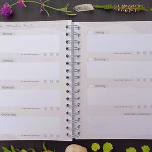 May include: A white planner with a gray spiral binding. The planner has a weekly layout with spaces for each day of the week, including Monday, Tuesday, Wednesday, Thursday, Friday, Saturday, and Sunday. The planner also has a section for notes and thoughts.
