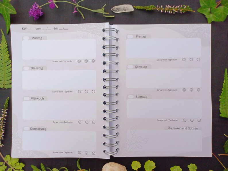 May include: A white planner with a gray spiral binding. The planner has a weekly layout with spaces for each day of the week, including Monday, Tuesday, Wednesday, Thursday, Friday, Saturday, and Sunday. The planner also has a section for notes and thoughts.