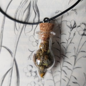 Könnte beinhalten: Eine Halskette mit einem Glasfläschchen mit Korkverschluss, gefüllt mit getrockneten Kräutern und Blumen. Die Kette ist an einem schwarzen Band befestigt und hängt vor einem weißen Hintergrund mit einem Blumenmuster.