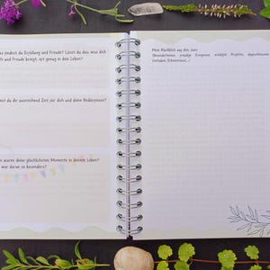 May include: A white spiral bound journal with a green cover. The cover has a wavy edge and features text in German. The text reads: "In was findest du Erp&uuml;llung und Freude? L&auml;sst du das, was dich erf&uuml;llt und Freude bringt, oft genug in dein Leben?"  "Nemst du dir ausreichend Zeit f&uuml;r dich und deine Bed&uuml;rfnisse?"  "Wann waren deine gl&uuml;cklichsten Momente in deinem Leben? Was war daran so besonders?"  "Mein R&uuml;ckblick auf den Juni:  Beziehungen, Freude, Errungenschaften, wichtige Projekte, abgeschlossene Vorhaben, Erkenntnisse..."
