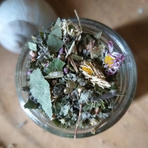 May include: A glass jar filled with dried herbs and flowers, including chamomile, lavender, and other botanicals. The jar is on a light brown surface.