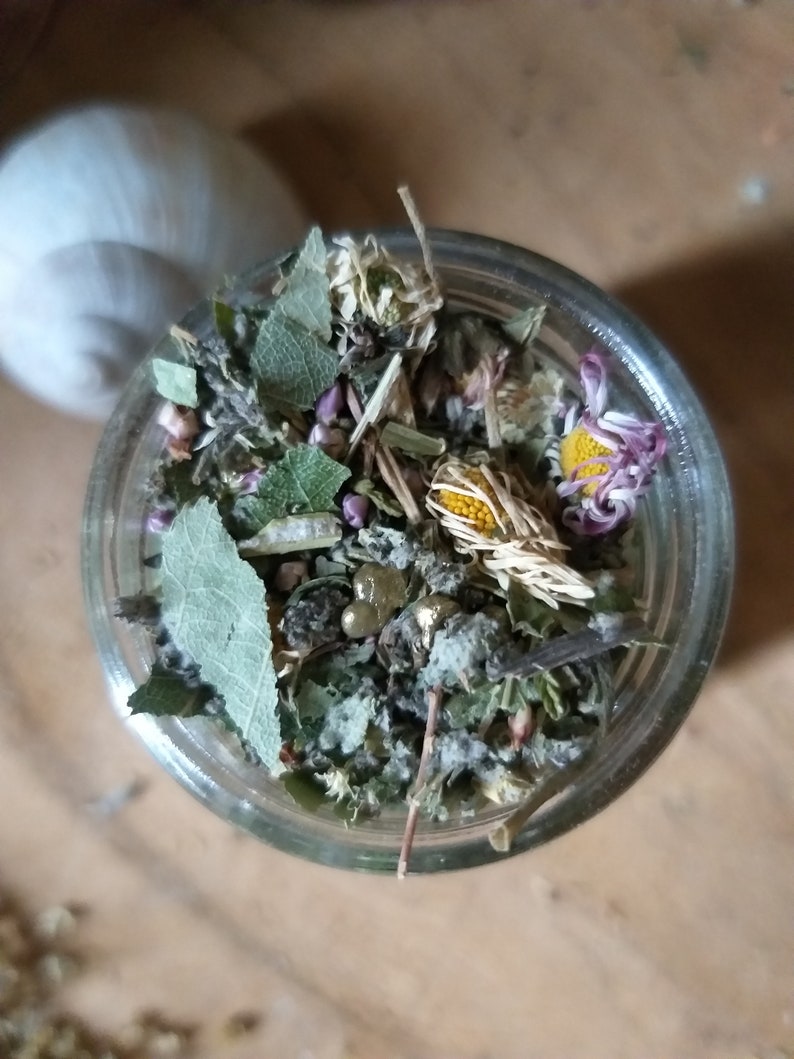 May include: A glass jar filled with dried herbs and flowers, including chamomile, lavender, and other botanicals. The jar is on a light brown surface.