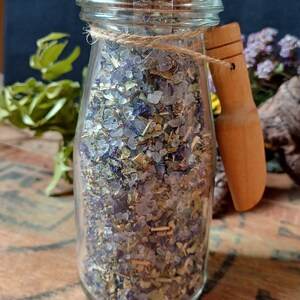 May include: A glass jar with a cork lid and a wooden handle filled with a mixture of dried herbs, flowers, and salt. The jar is tied with twine and sits on a wooden surface.
