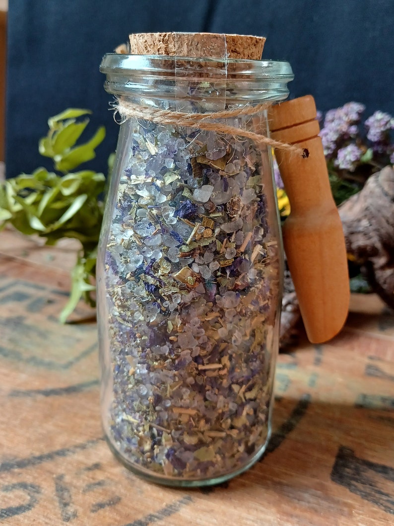 May include: A glass jar with a cork lid and a wooden handle filled with a mixture of dried herbs, flowers, and salt. The jar is tied with twine and sits on a wooden surface.