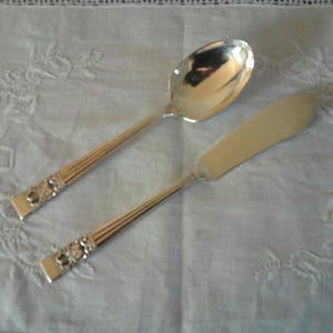 May include: A silver-toned metal spoon and a flat-bladed knife. Both utensils feature a polished finish, a handle with three vertical ridges, and a decorative rectangular section with embossed floral detailing near the base of the handle.