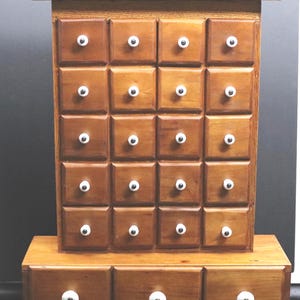 May include: A wooden apothecary chest featuring twenty-three small square drawers arranged in a grid. Each drawer is equipped with a small, round white knob with a black center. The unit has a flat top ledge and a wider base section with three drawers.